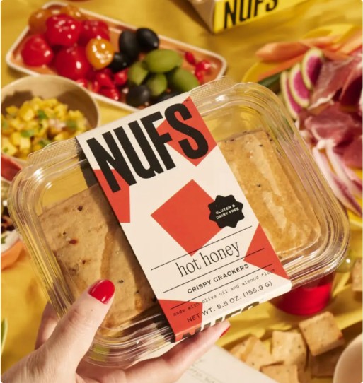 Hand holding a NUFS “hot honey” crispy crackers clamshell over a colorful snack spread.