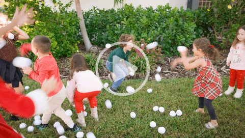 Children having a playful snowball fight on a green lawn, with smiles and holiday outfits in a festive outdoor setting.