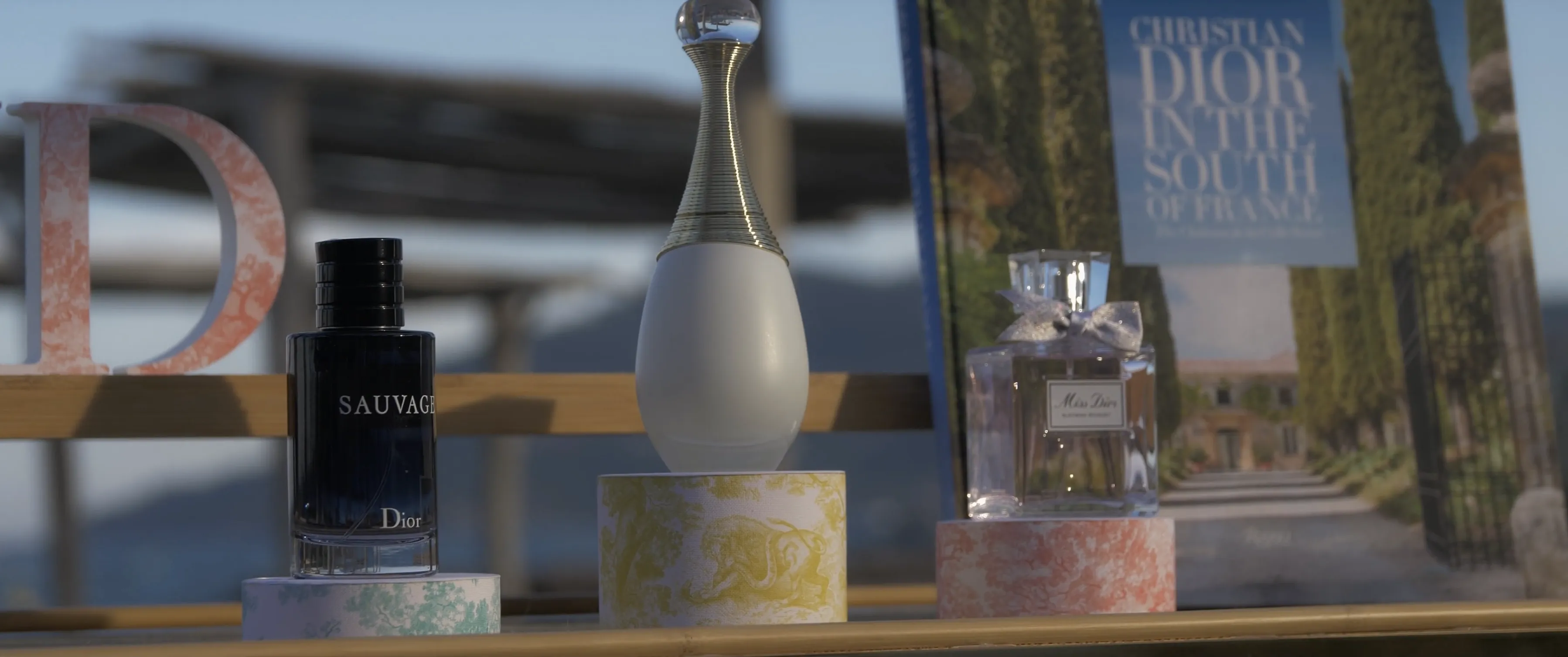 Three elegant perfume bottles, including Dior's "Sauvage" and "Miss Dior," are displayed on decorative pedestals with a backdrop of the "Christian Dior in the South of France" book under soft lighting.