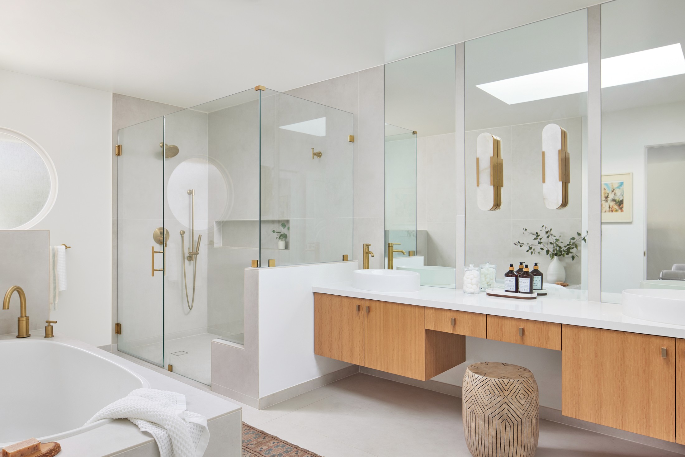 Stunning master bathroom complete with a white quartz countertop, mounted stone light fixtures, bronze fixtures, vertical grained flat panel cabinetry, large white porcelain tile surrounds, a skylight, built-in vanity space, and large soaking tub crafted by Jubilee Interiors in Los Angeles, California