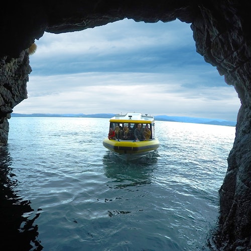 Жълта лодка с хора вътре се вижда от пещера, плаваща по спокойна синя вода с облачно небе на заден план.