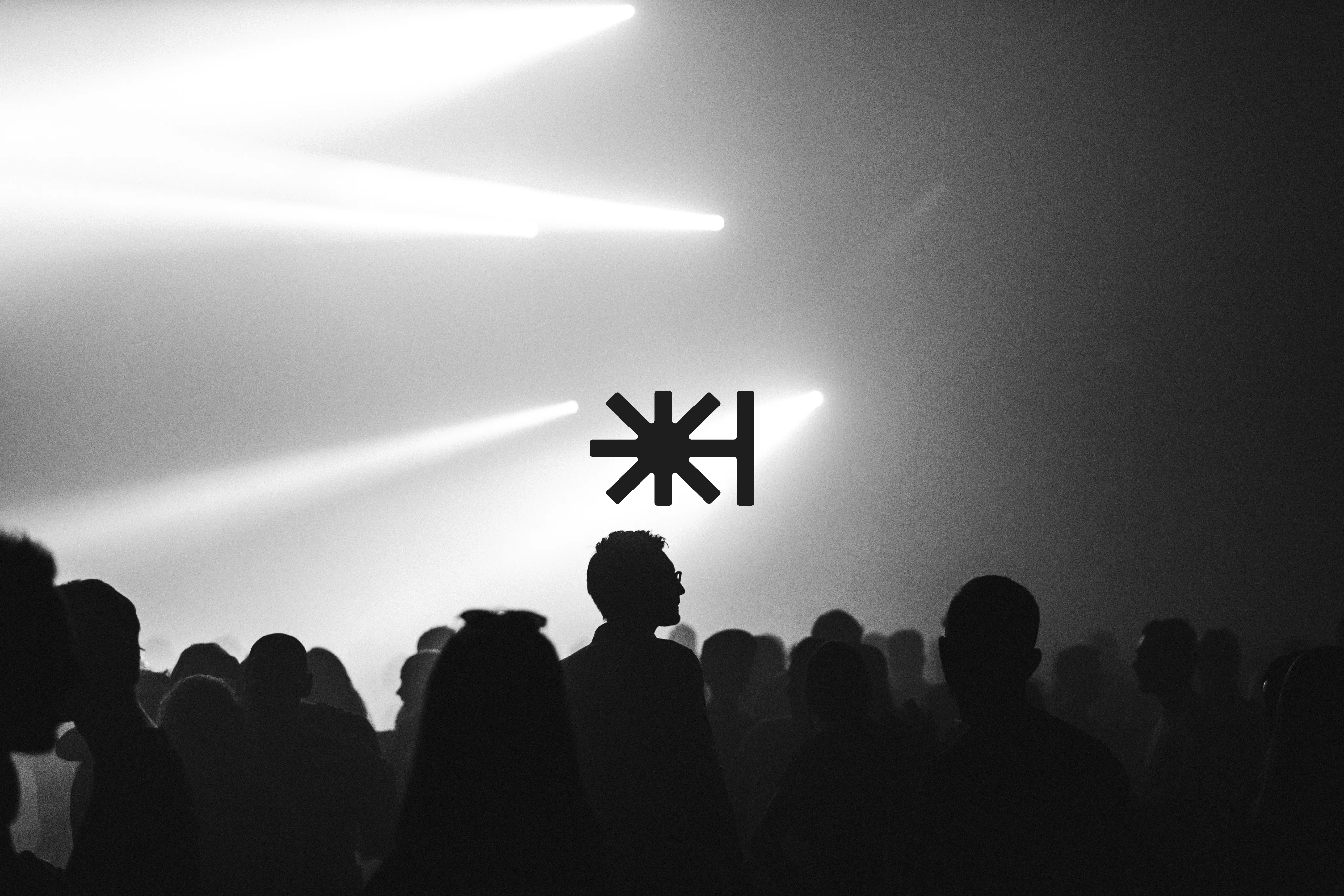 Silhouettes of people at a concert under beams of light, with a graphic symbol in the center. Black and white image.