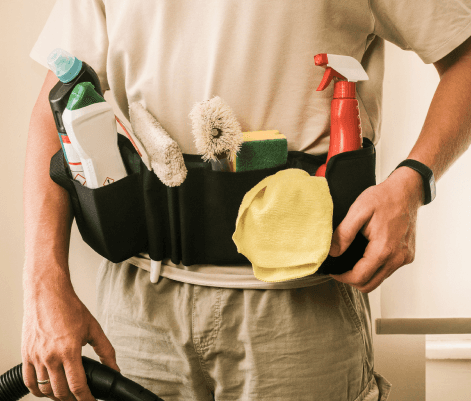 A person is wearing a utility belt filled with various cleaning supplies, including spray bottles, sponges, and brushes. The individual is holding a vacuum cleaner hose in one hand. The scene suggests a cleaning or maintenance task, with a focus on the organized tools for the job.