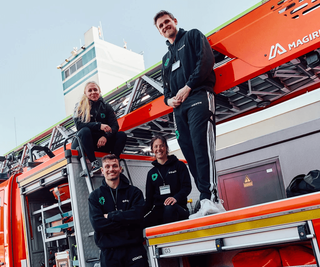 Zwei junge Frauen und zwei junge Männer in Strong Partners Sportkleidung sitzen und stehen auf einem Feuerwehrfahrzeug