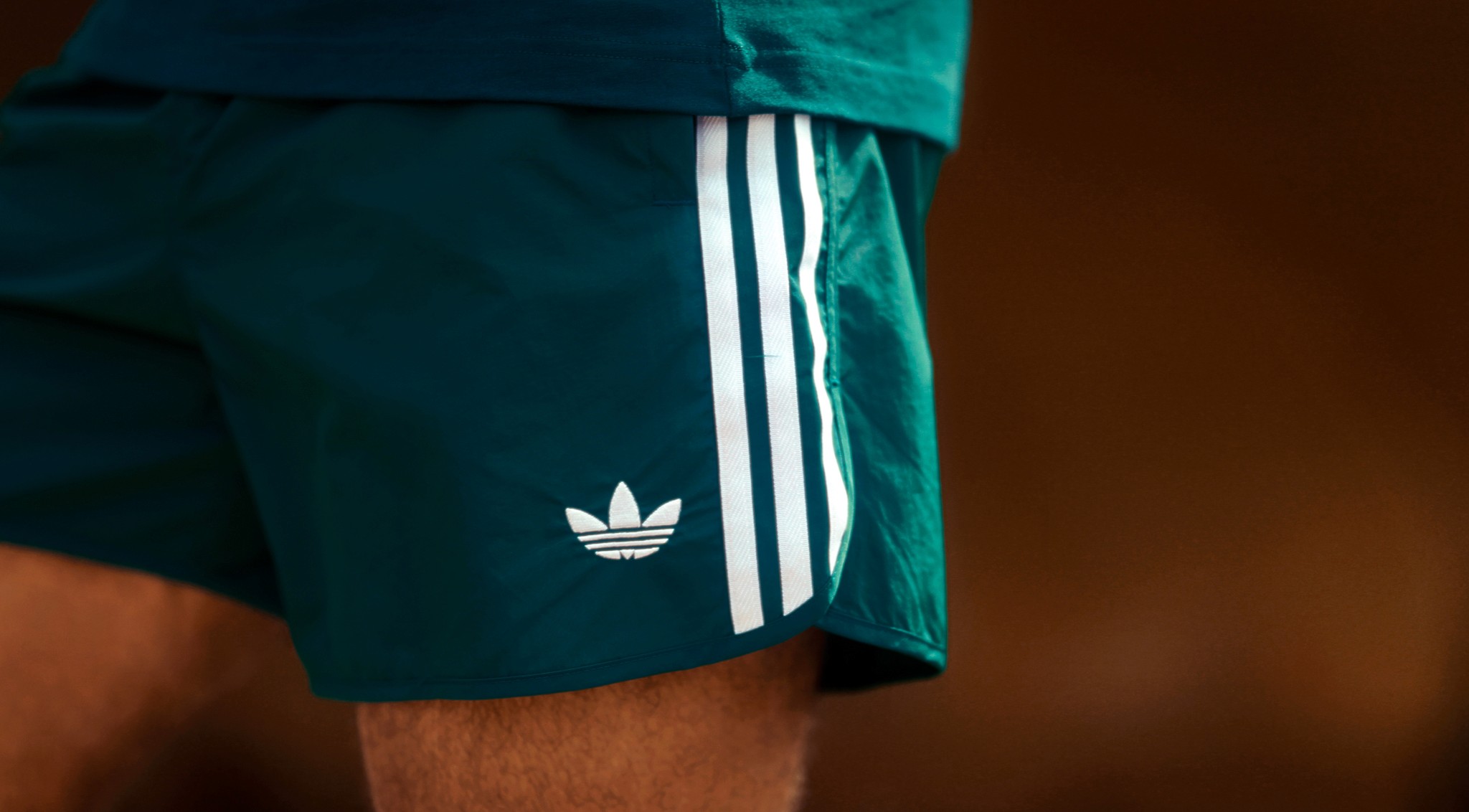 A person wearing green athletic shorts with white stripes stands outdoors, showcasing the classic Adidas logo against a blurred brown background.