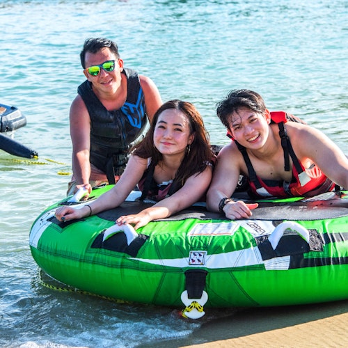 三个人穿着救生衣和泳装，微笑着坐在一个绿色的充气船上，浅浅清澈的水中。