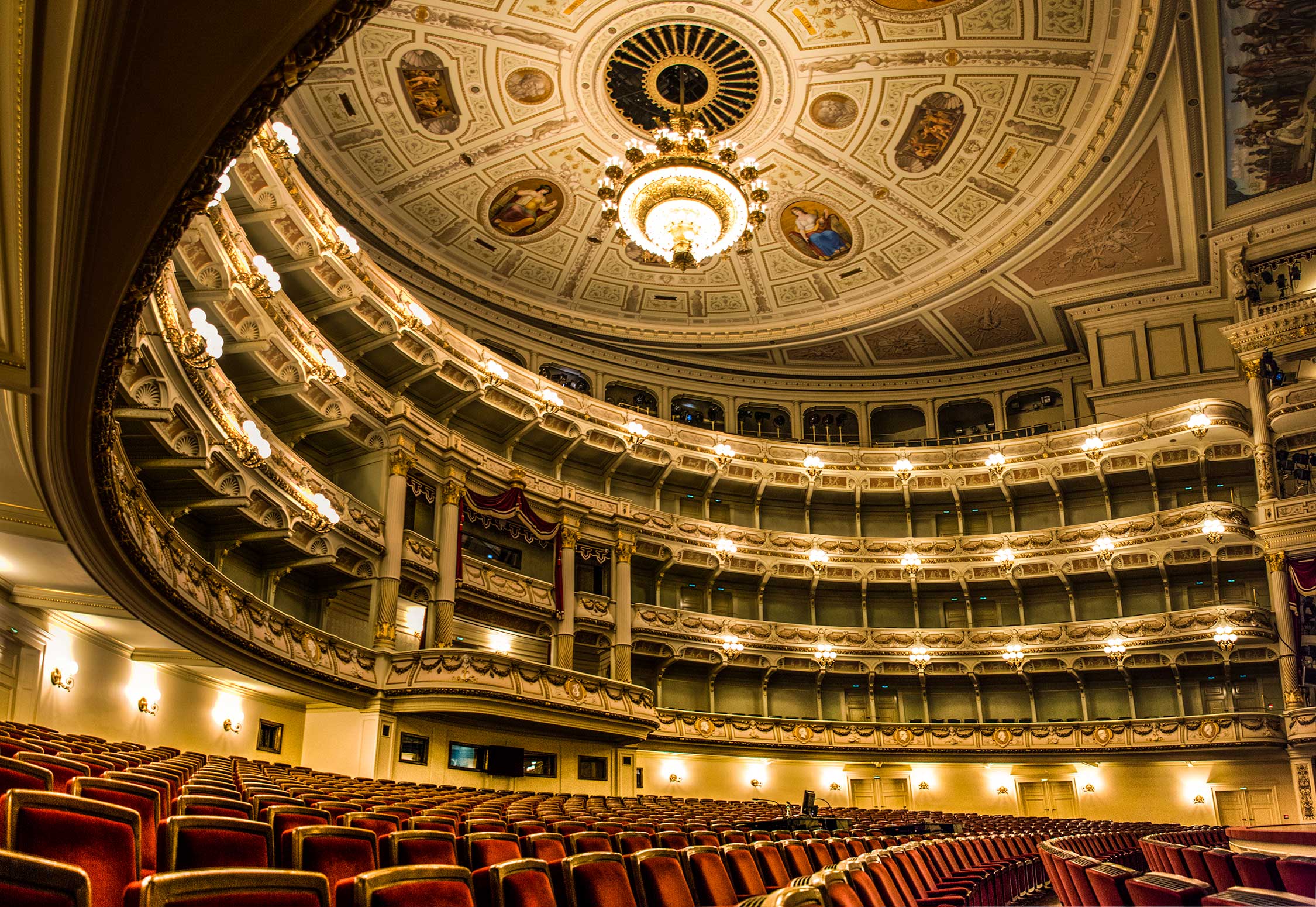 Interior view of a grand opera house with gold-trimmed balconies, red seating, and an ornate domed ceiling.
