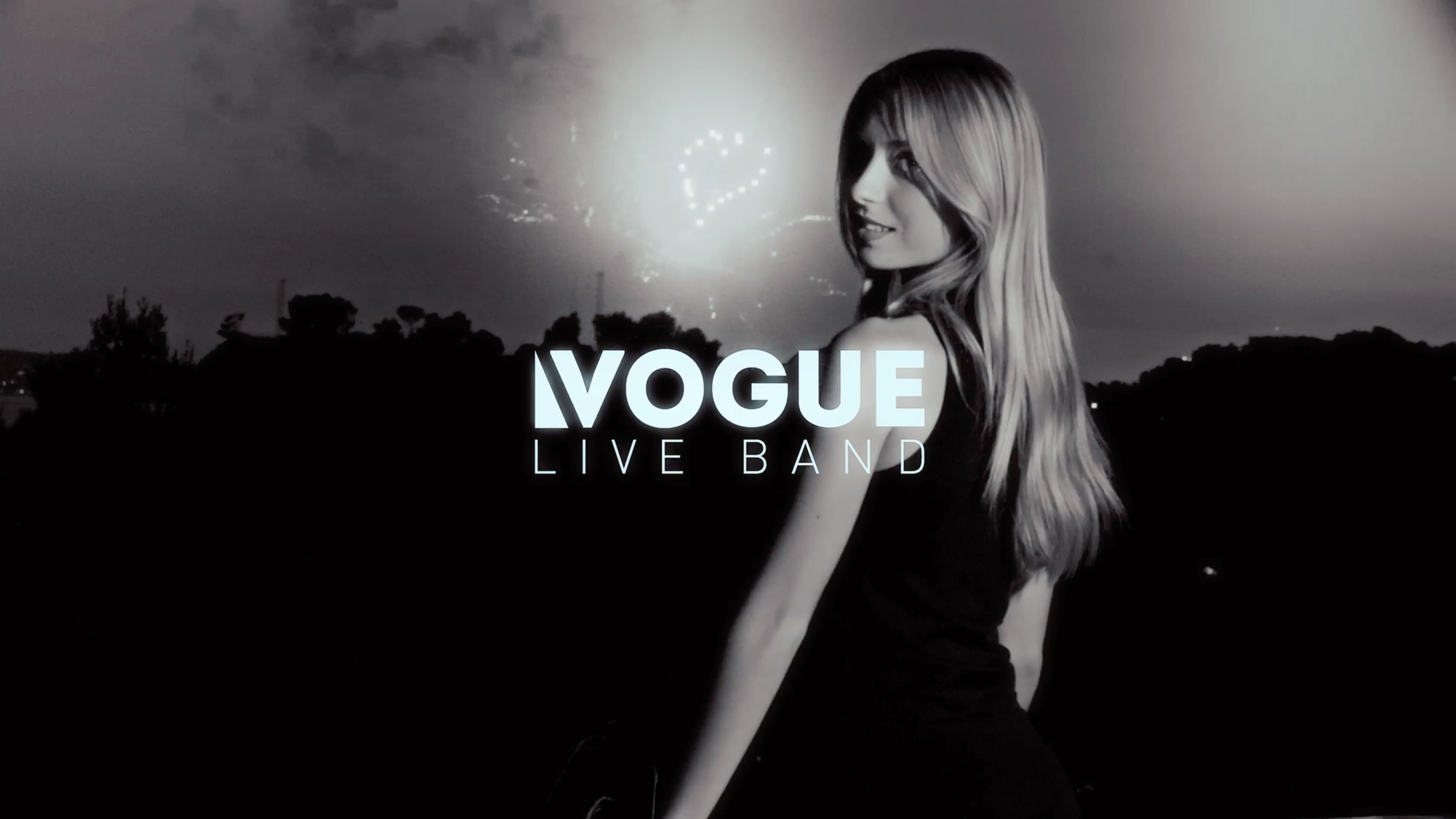 A black and white image featuring a woman in a dark dress with long hair, standing against a nighttime backdrop with fireworks, alongside the text "Vogue Live Band."