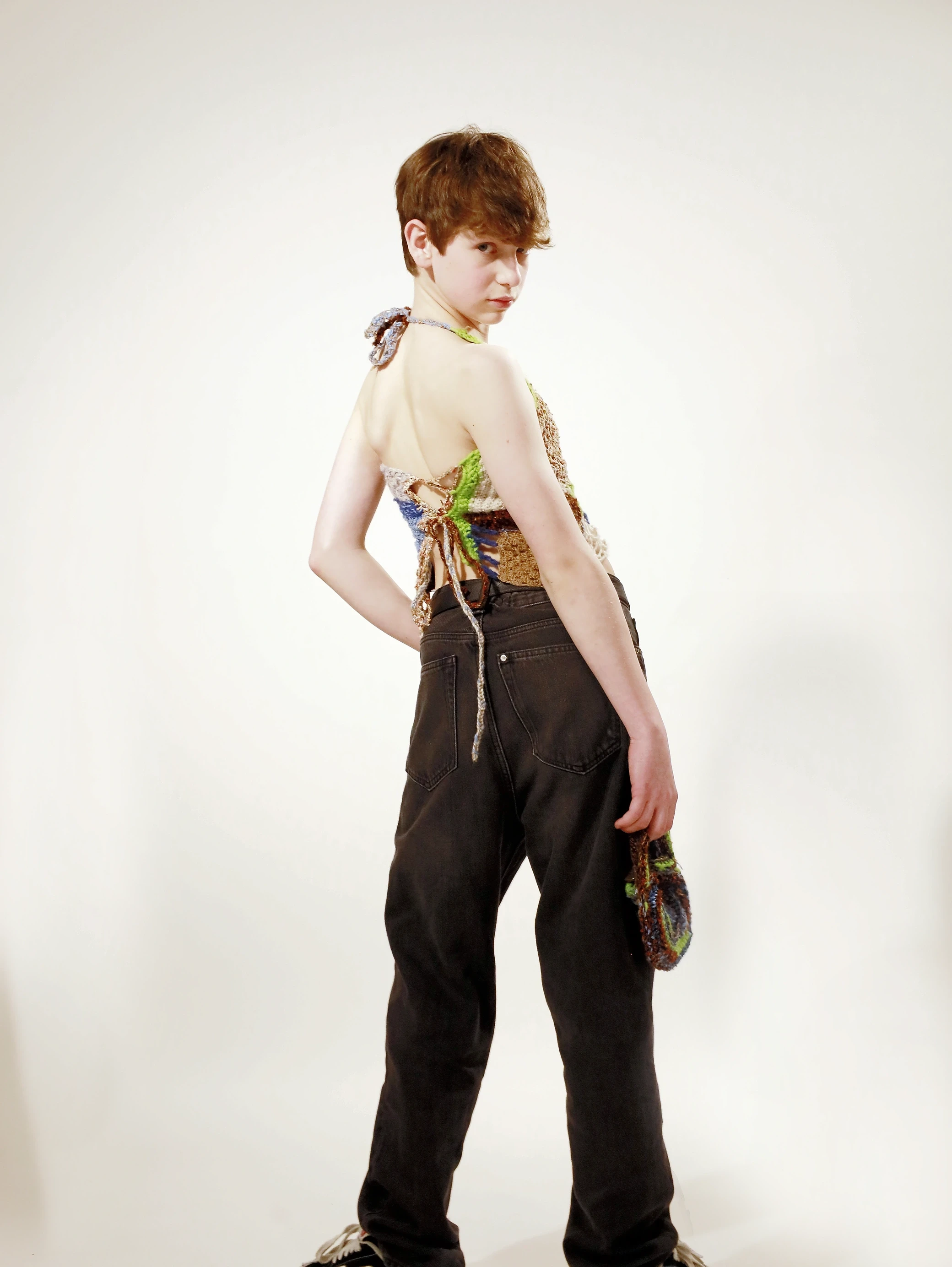 A person with short hair poses in stylish clothing, looking back over their shoulder against a plain background.