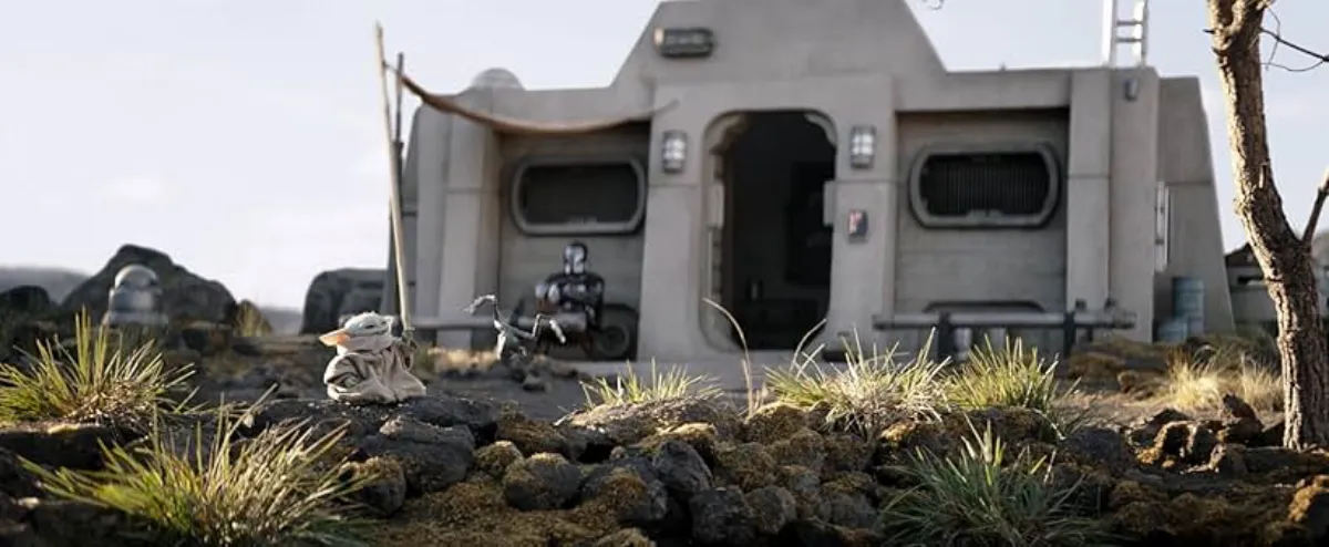 Grogu sits in a field of rocks and grass, seemingly peaceful as The Mandalorian sits with his feet up in front of their home.