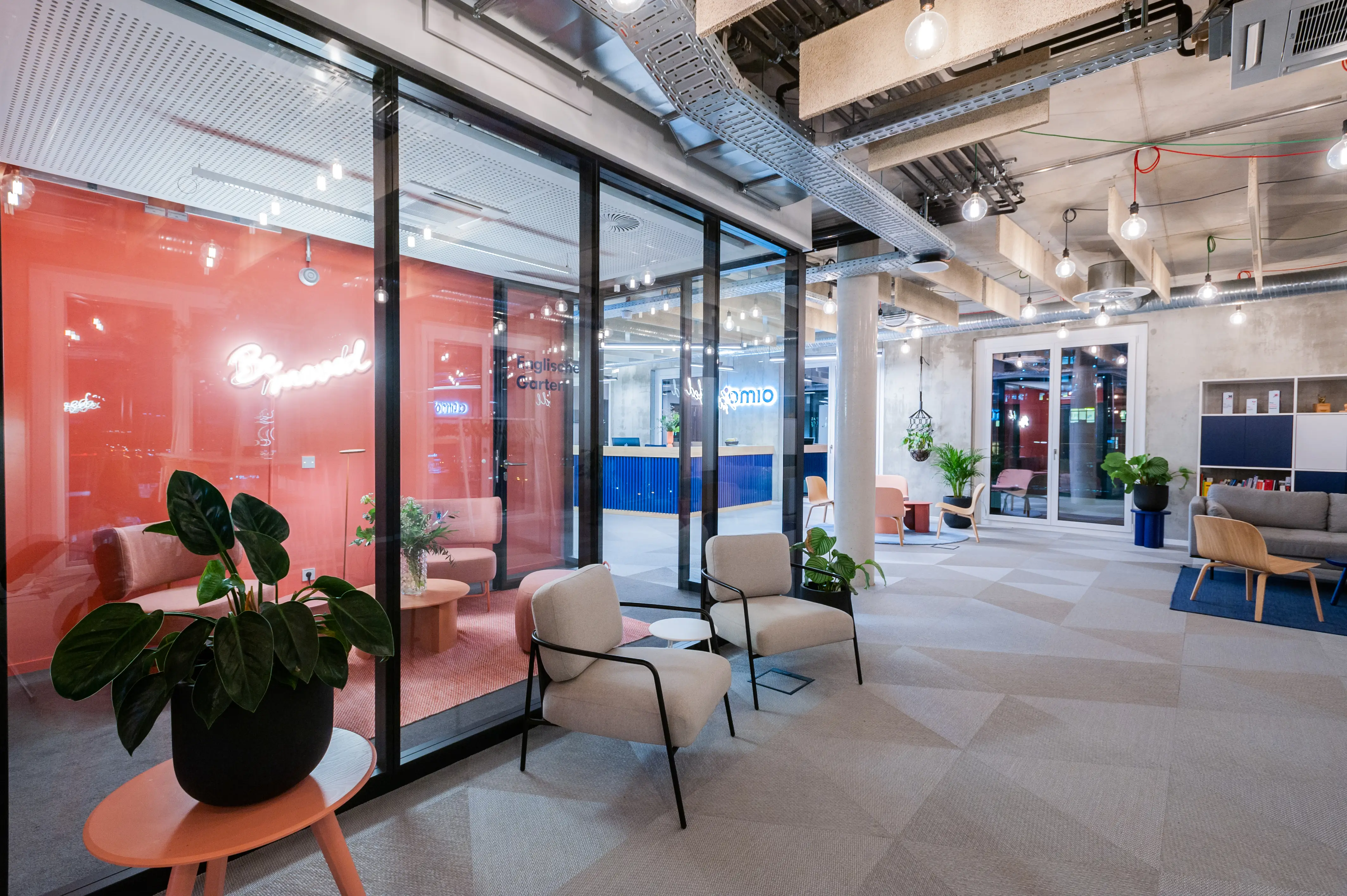 Corridor at OMIO along a glazed meeting room with coral-red interior and a neon script sign, cream lounge chairs, a peach side table with a potted plant and exposed services on the concrete ceiling.