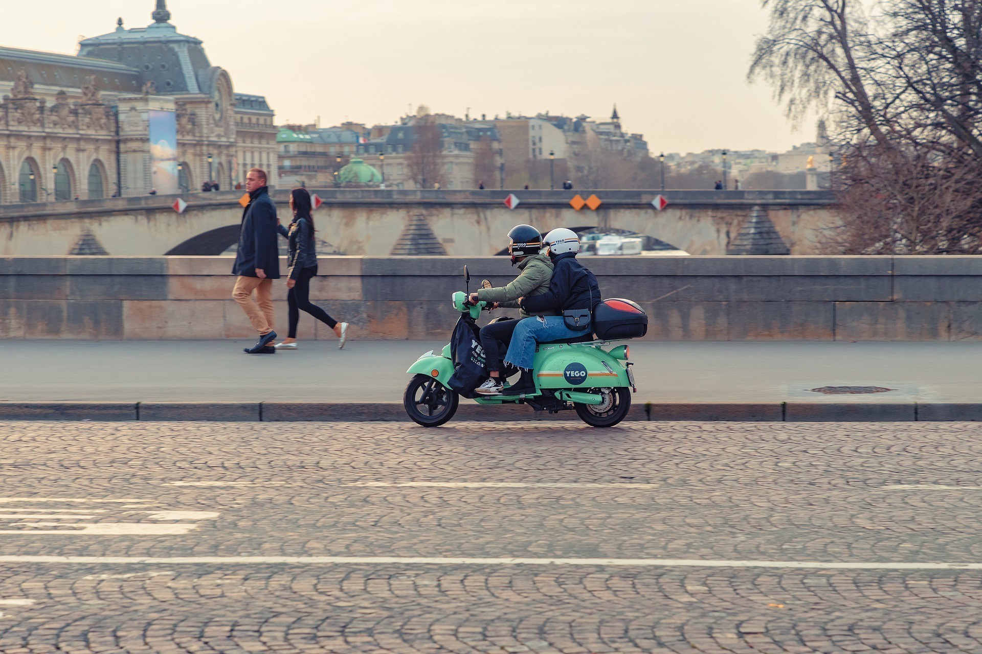 Een persoon rijdt op een groene scooter langs een pad, met voetgangers die in de buurt lopen en stadsgebouwen op de achtergrond.