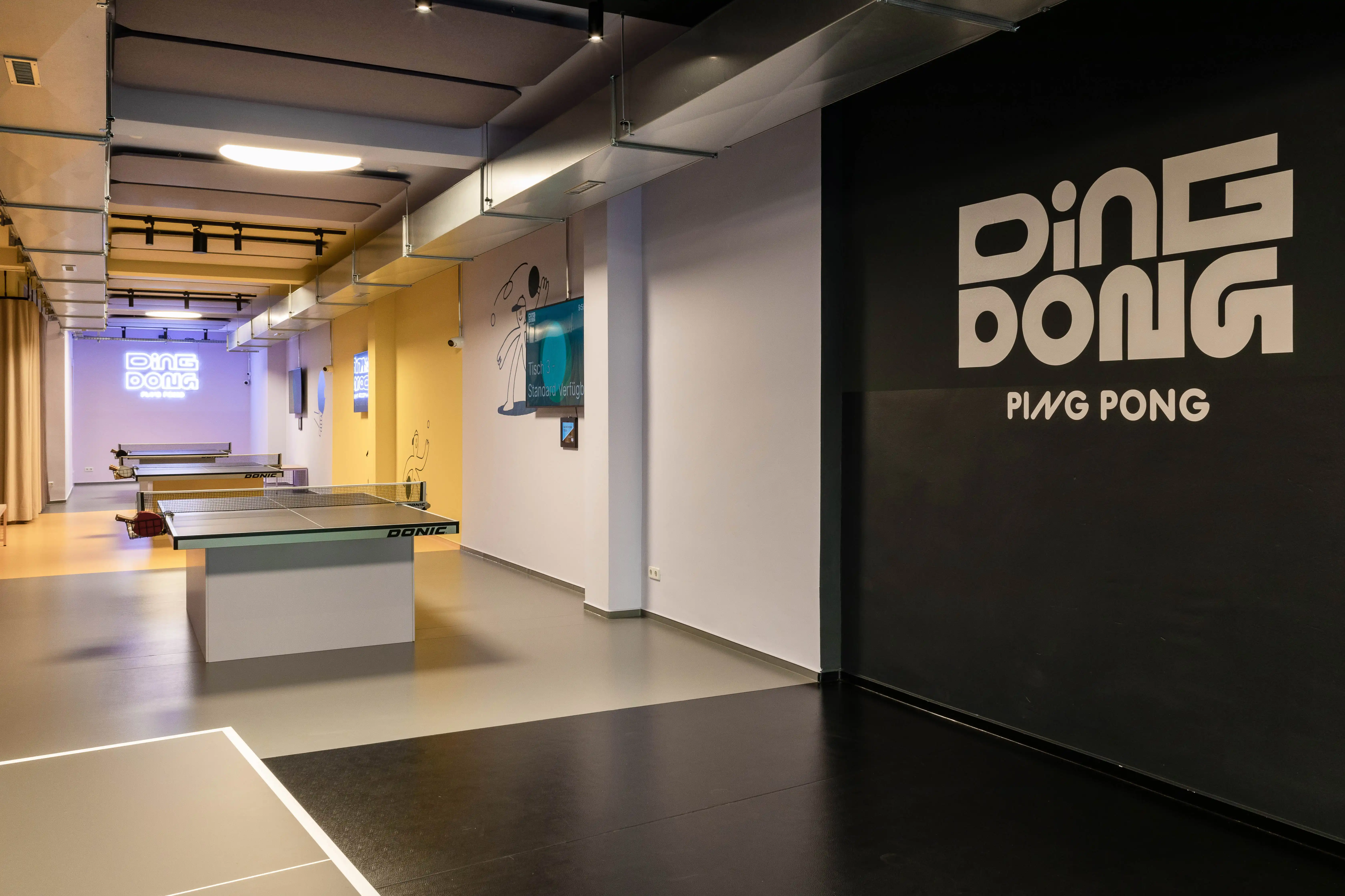Interior entrance of DING DONG PING PONG with a large white logo on a black wall and table tennis tables in the warm yellow hall behind.