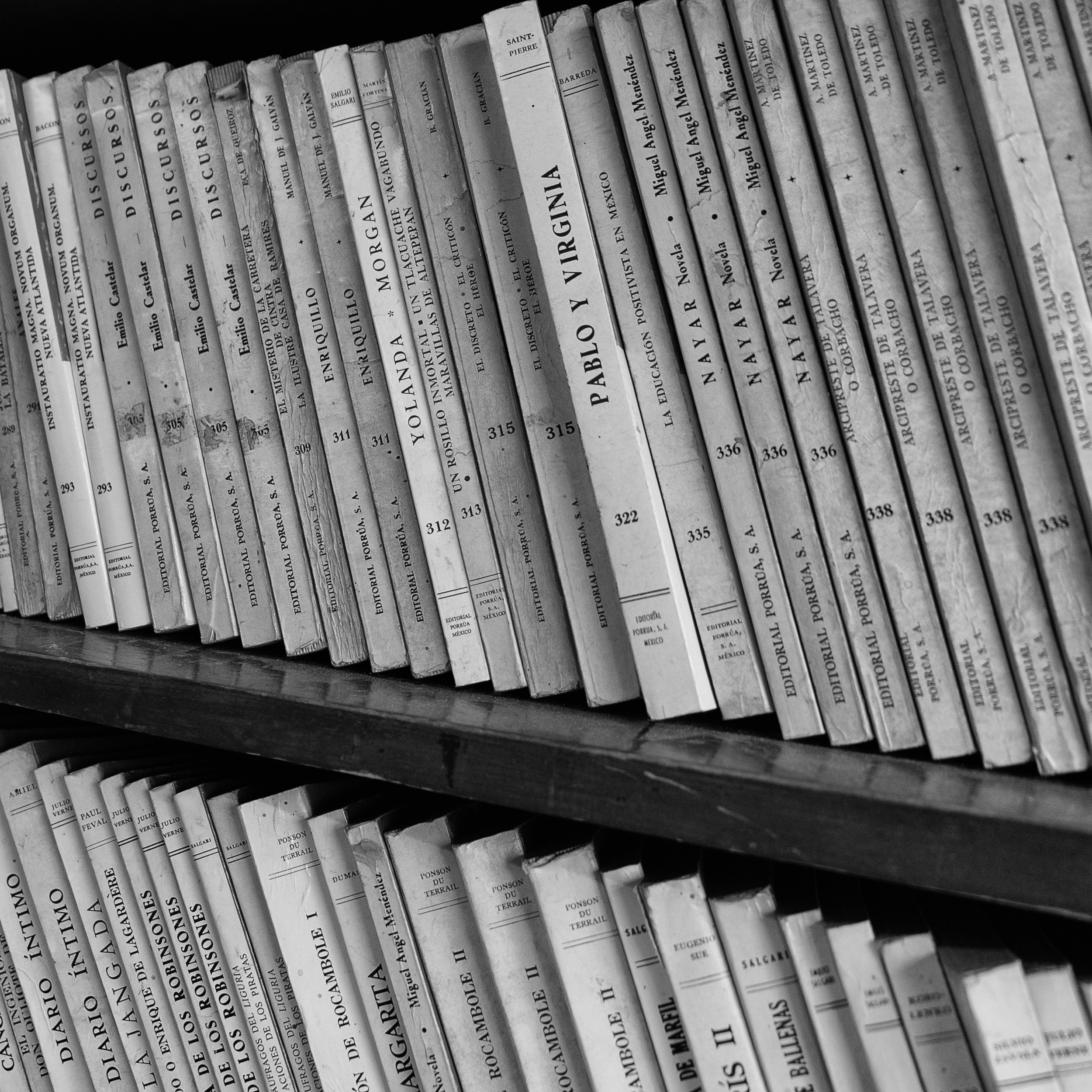 Black and white close-up of a bookshelf filled with vintage paperback books in Spanish, their spines showing signs of age and wear.