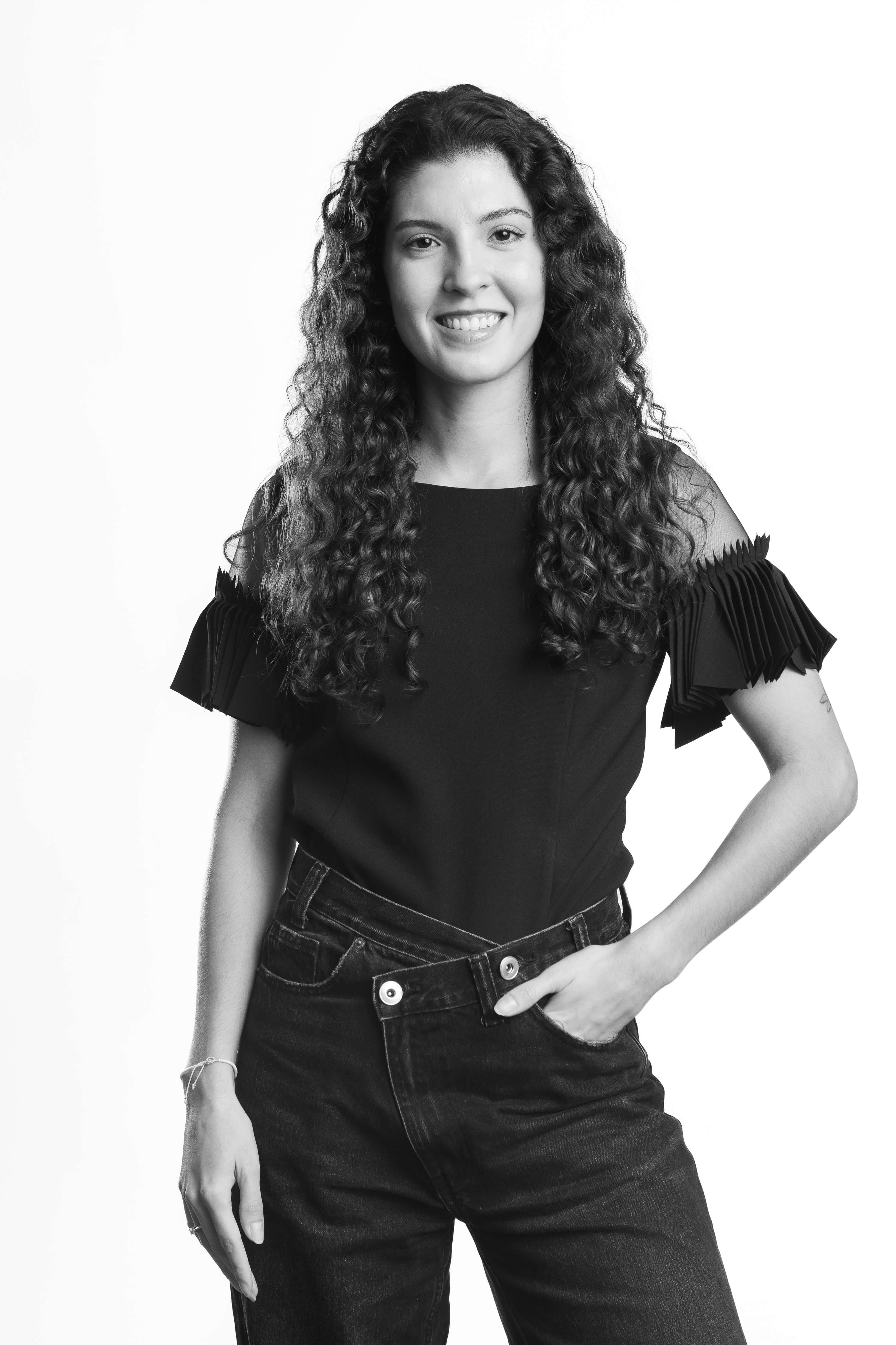 Black and white portrait of a smiling woman with long curly hair, wearing a black top with pleated sleeves and dark jeans. She stands confidently with one hand in her pocket.