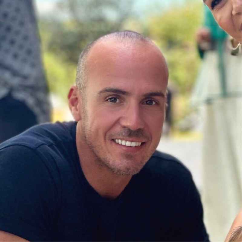 A smiling person wearing glasses and a patterned shirt stands against a plain white background.