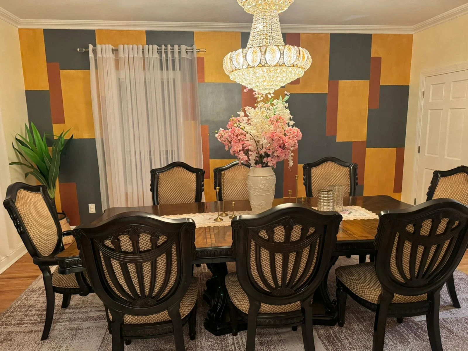 Elegant dining room with a bold geometric accent wall in gold, charcoal, and rust tones, featuring a crystal chandelier, traditional dark wood furniture, and a large floral centerpiece.