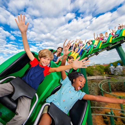 Children with hands up, excitedly riding a green rollercoaster. Blue sky with scattered clouds in the background.