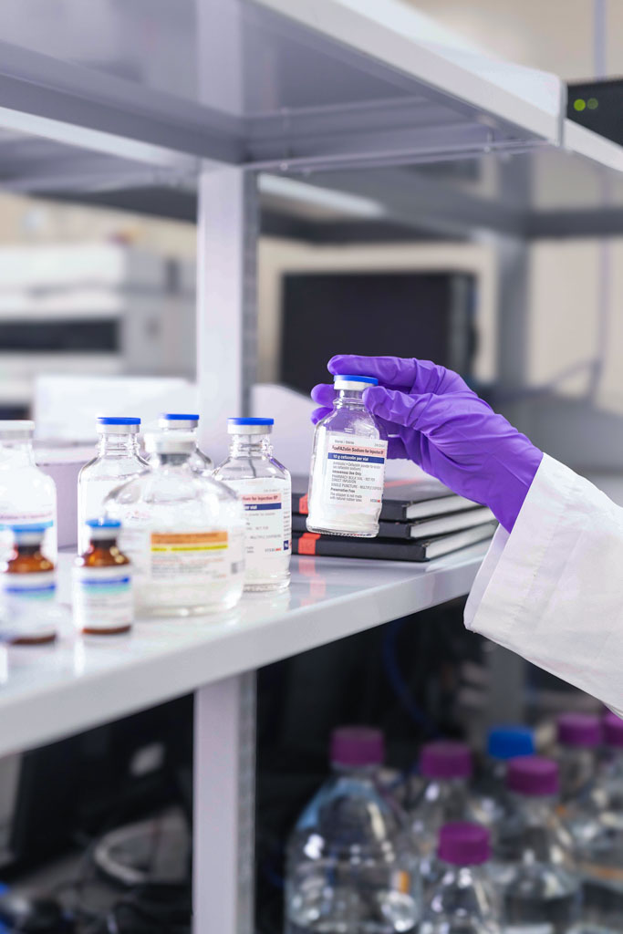 Gloved hand holding a small glass vial on a laboratory shelf with other vials and bottles.