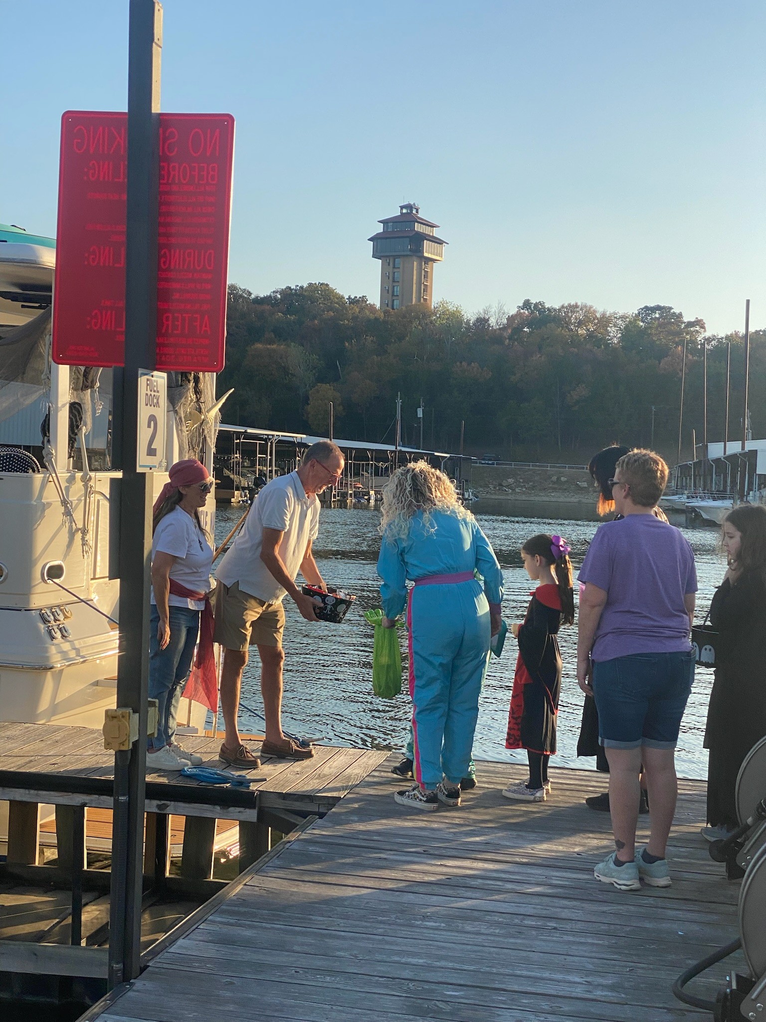 A group of people, including children in costumes, stand on a wooden dock next to a yacht with a backdrop of calm water, trees, and a tall structure, under a clear blue sky in the evening light.