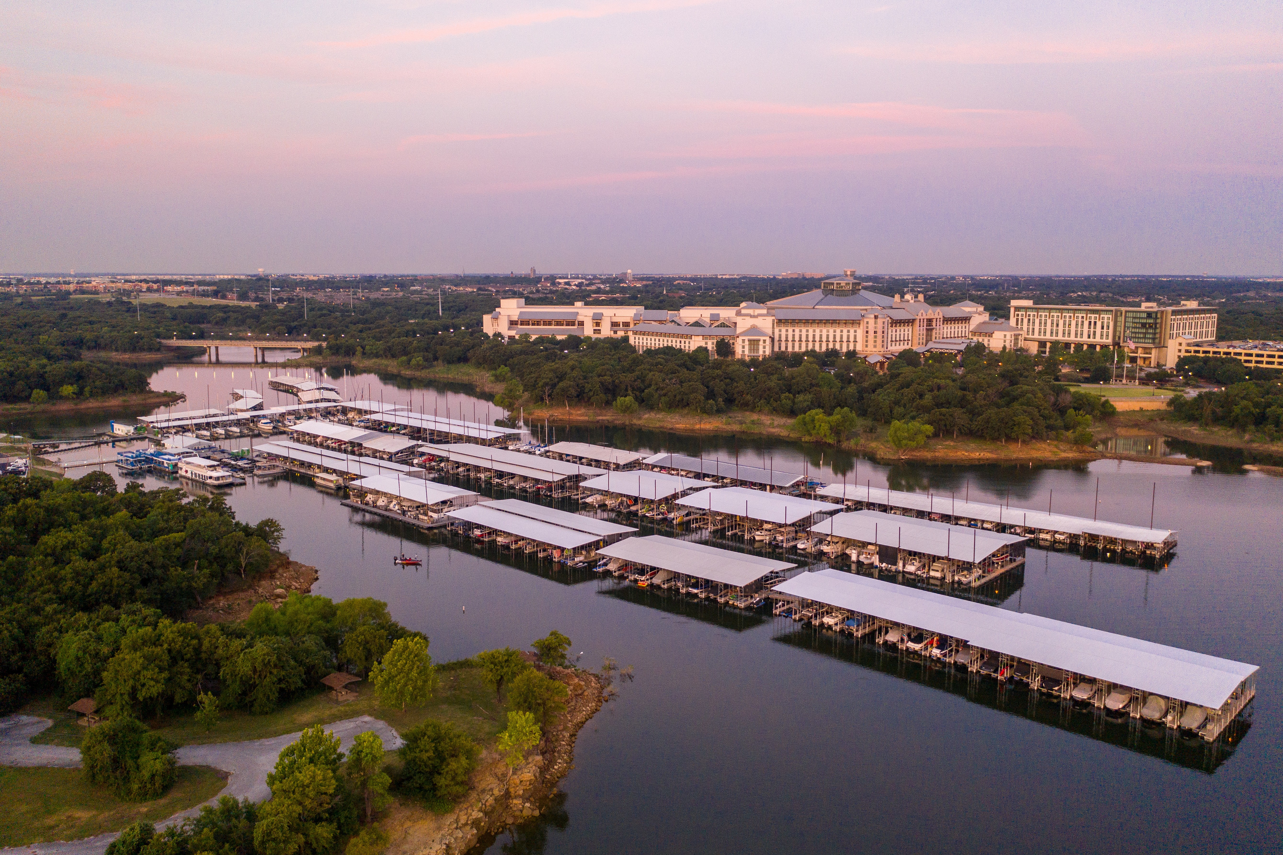 Aerial view of Silver Lake Marina with multiple boat docks extending into the tranquil water, surrounded by lush green trees and positioned against the backdrop of expansive, architecturally striking buildings under a soft, pastel sky at dusk, capturing the serene ambiance of lakeside living.