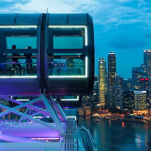 Two people inside a lighted observation pod on a Ferris wheel overlook a city skyline and waterfront at dusk.