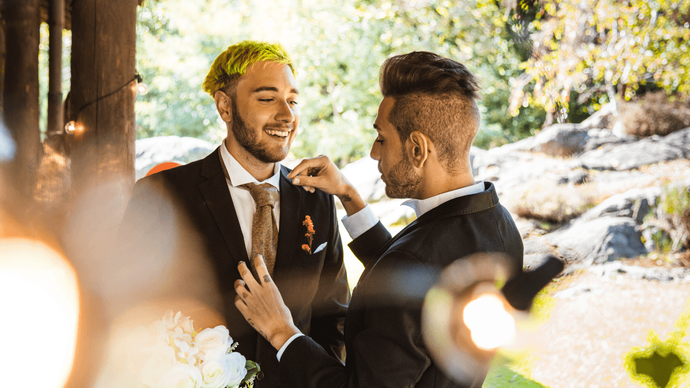 Two well-dressed men smile and pose joyfully together outdoors, surrounded by greenery.