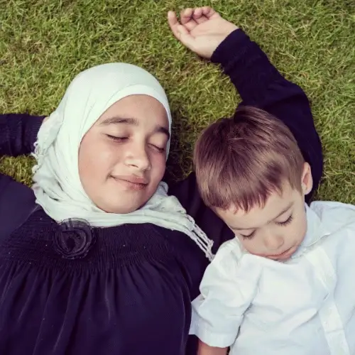 Young girl in a navy hijab resting on green lawn beside her little brother, representing family post-procedure care in Melbourne.
