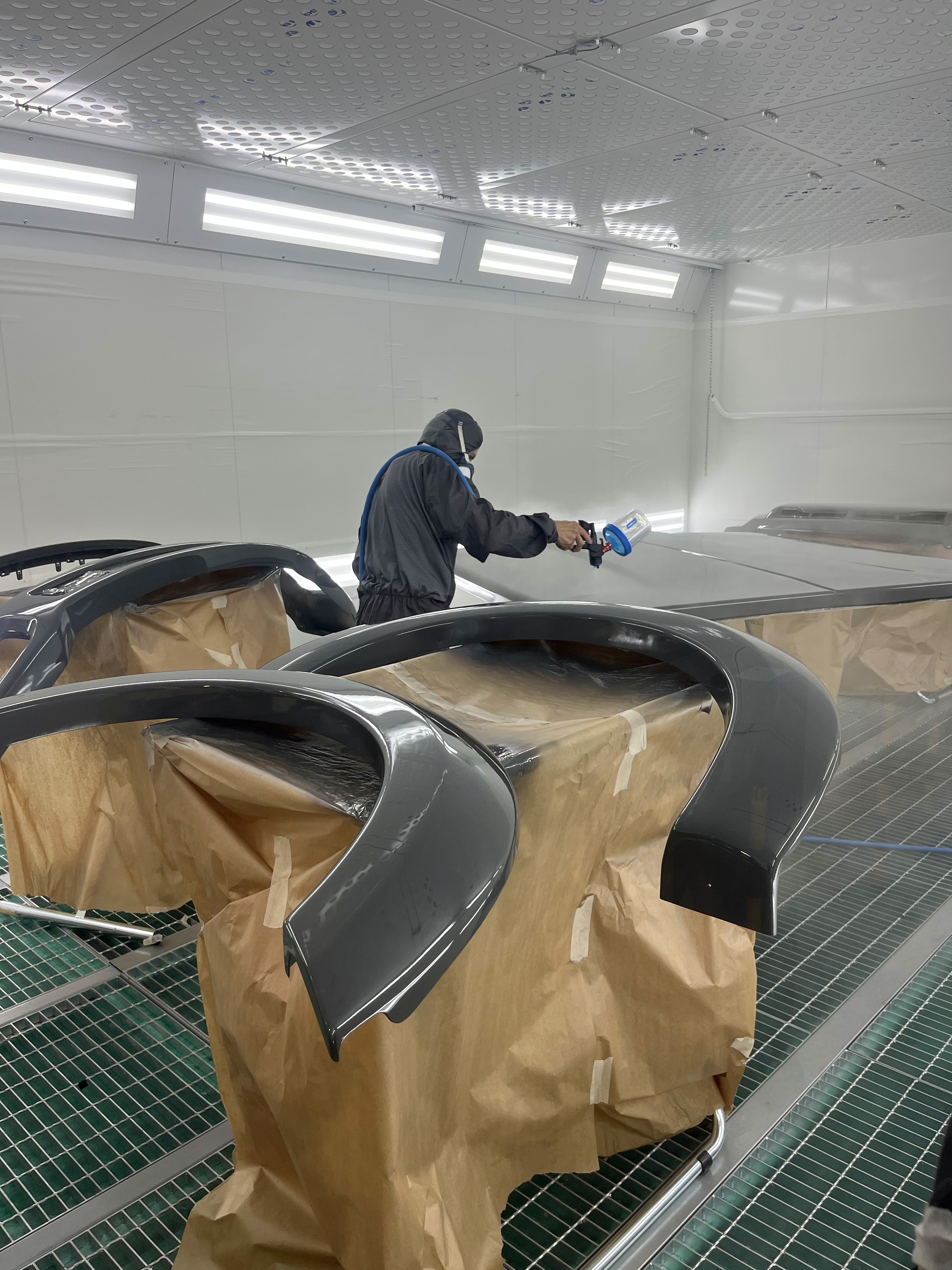 Two people in a workshop, one standing and smiling while the other leans over a car, working on it.