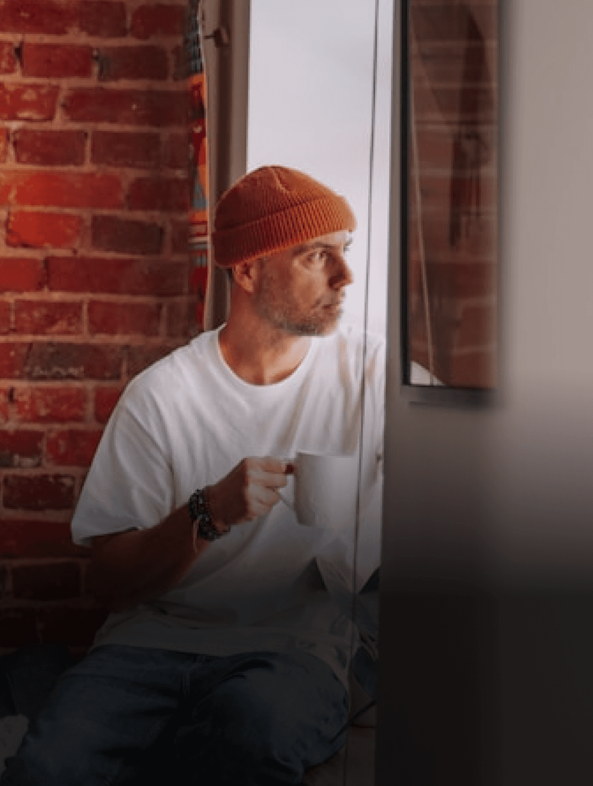Photograph of a man wearing a beanie and holding a mug, sitting on the ledge of a window and staring outside