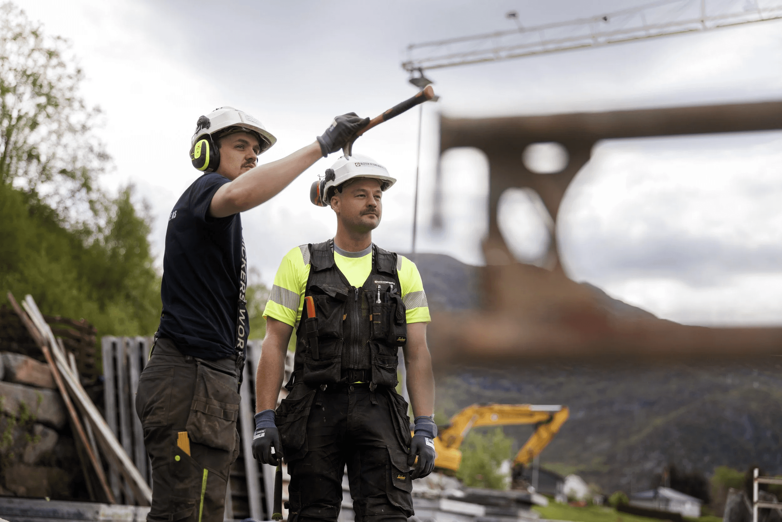 Two construction workers in safety gear stand on a site with a bridge structure in the background, discussing.