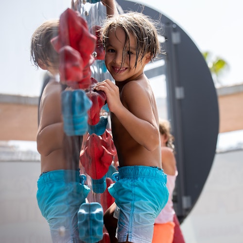 一个穿着蓝色短裤的孩子在游乐场的攀爬架上，面向相机微笑，背景中还有另一个孩子。