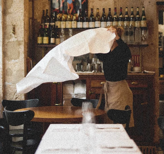 Waiter unfolding a white tablecloth in a cozy restaurant filled with wine bottles and wooden details, capturing a candid moment of daily ritual.