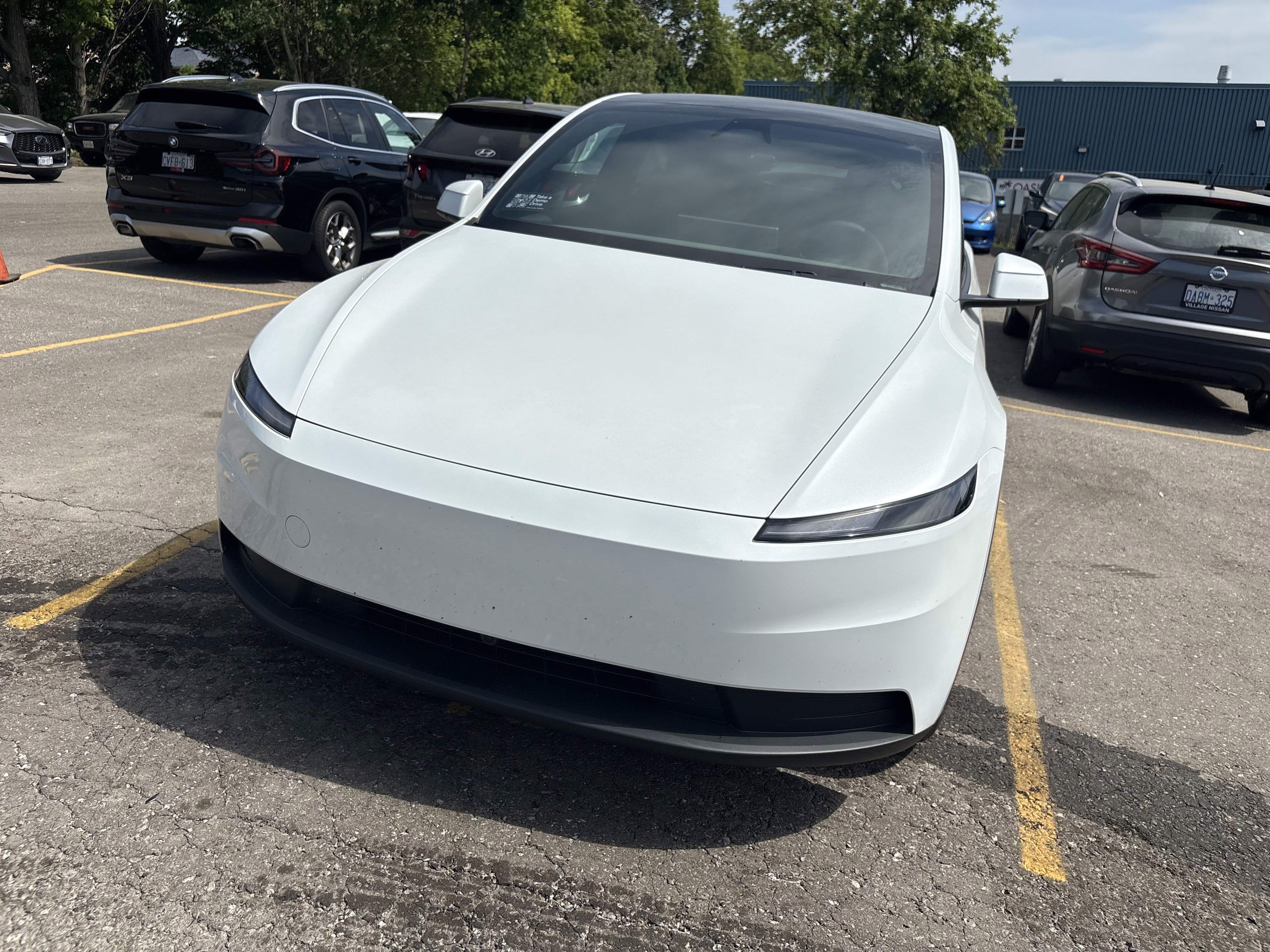 A sleek, white Tesla Model Y electric car is parked in a parking lot, surrounded by several other vehicles, under a clear blue sky.