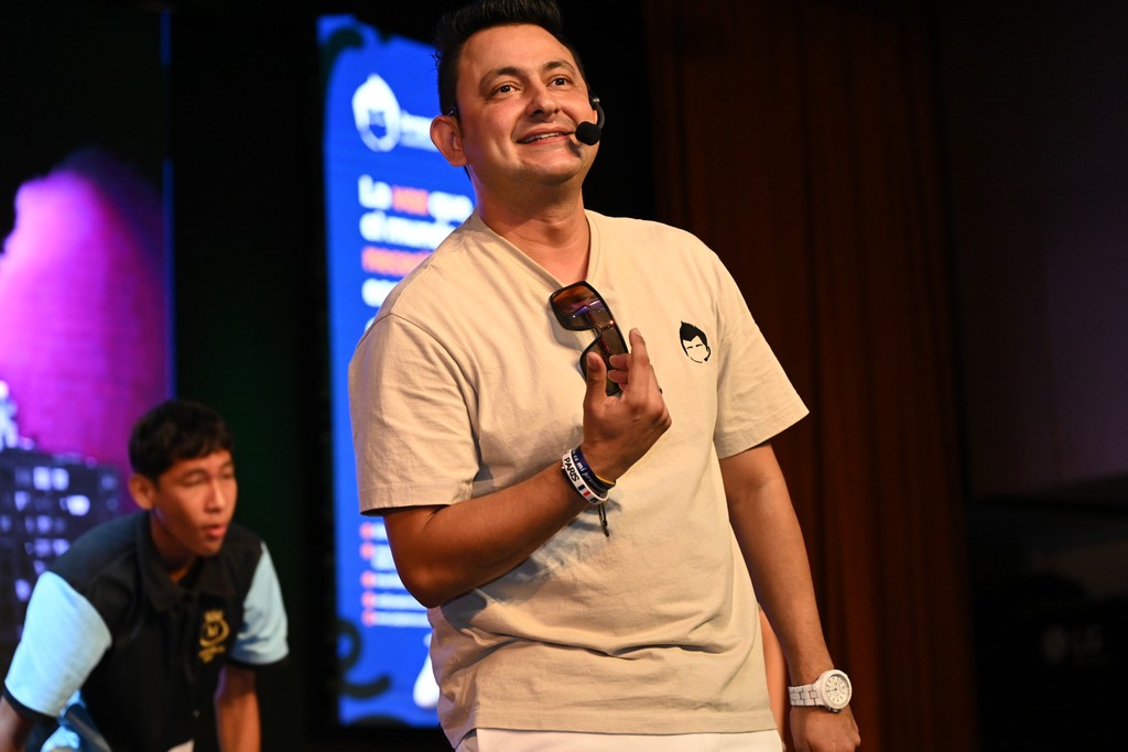 Jorge Baena sonriendo en el escenario, sosteniendo unas gafas de sol durante una conferencia frente a jóvenes asistentes.