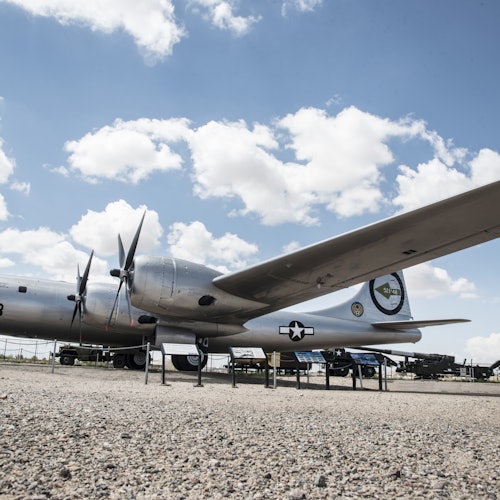 一架銀色的第二次世界大戰時期飛機在室外展示，配有螺旋槳和軍事標記，天上有些雲。