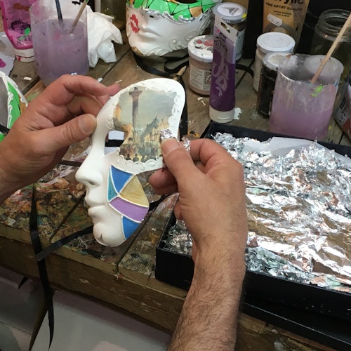 Mani che tengono una maschera decorata sopra un piano di lavoro ingombro di materiali per dipingere, pennelli e fogli di alluminio.