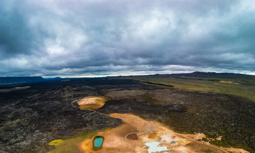 Aerial view of the Krafla lava fields at Leirhnjúkur in Iceland, featuring expansive black basalt lava flows, geothermal activity, volcanic craters, and rugged terrain beneath dramatic storm clouds.