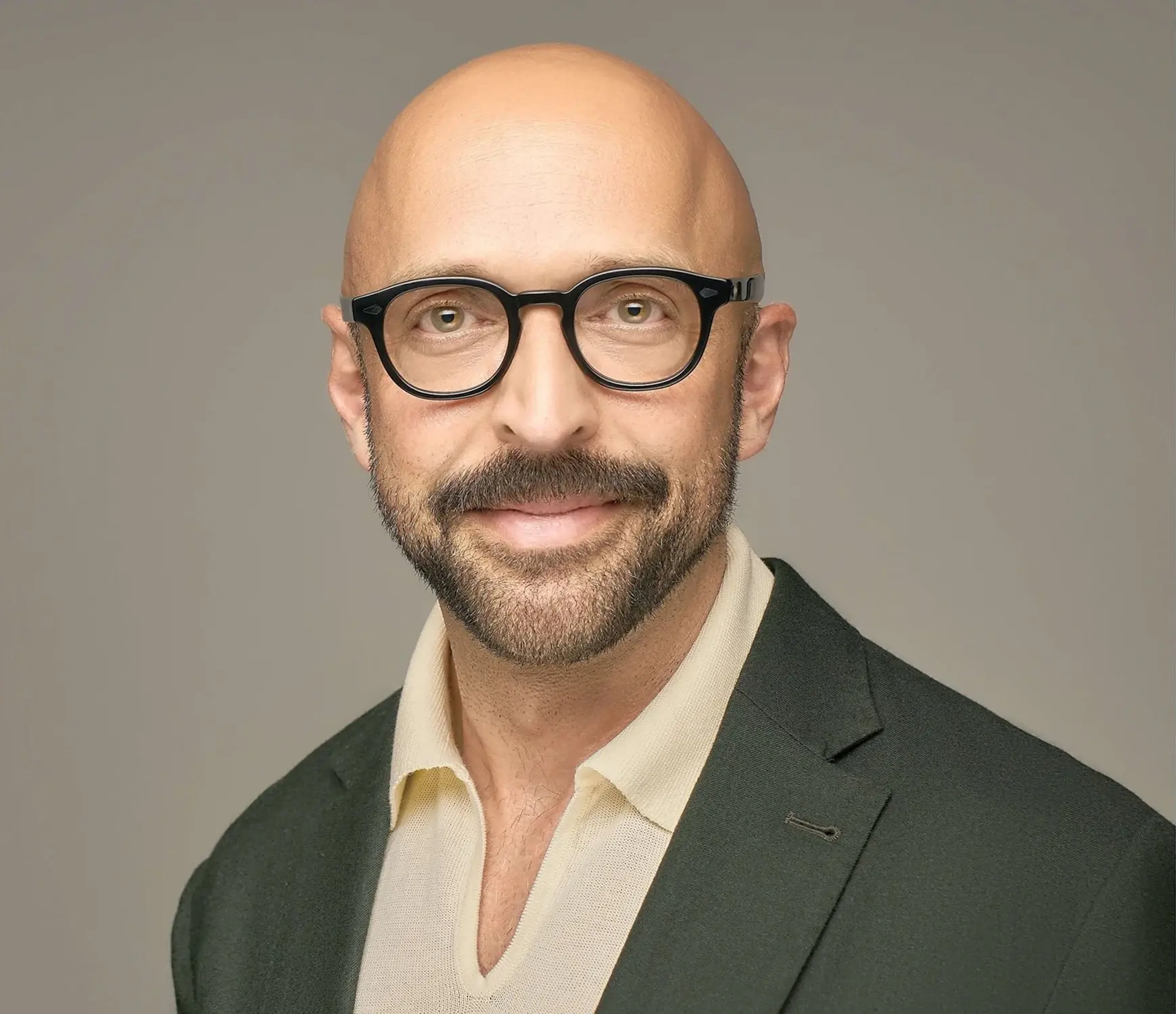 A bald, bearded man wearing round black glasses and a dark green suit jacket over a light-colored shirt smiles at the camera, set against a plain gray backdrop.