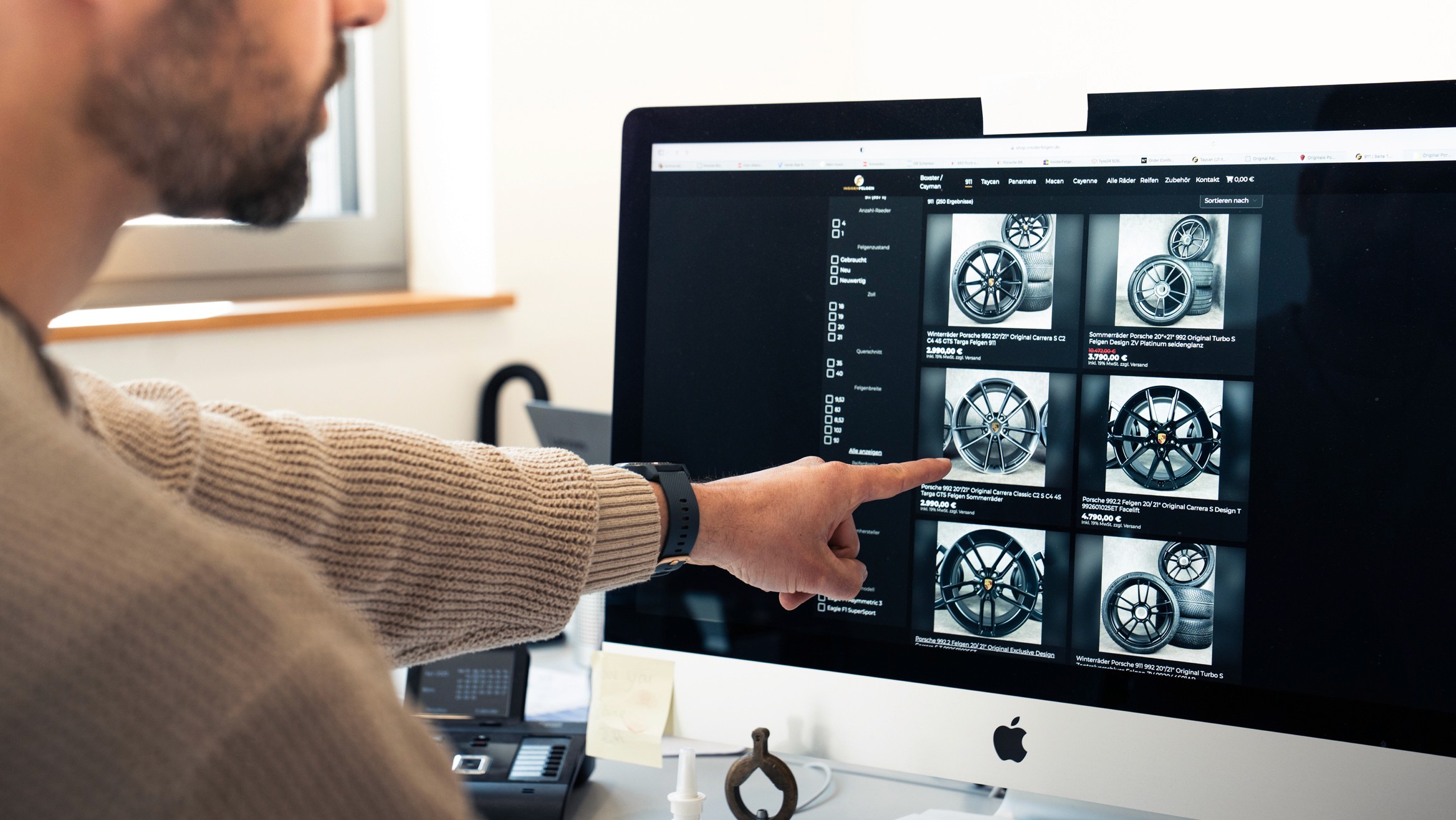 Mitarbeiter zeigt auf eine Auswahl an Porsche-Felgen im Onlineshop auf einem Monitor, Bildschirmansicht mit verschiedenen Felgendesigns und Preisen