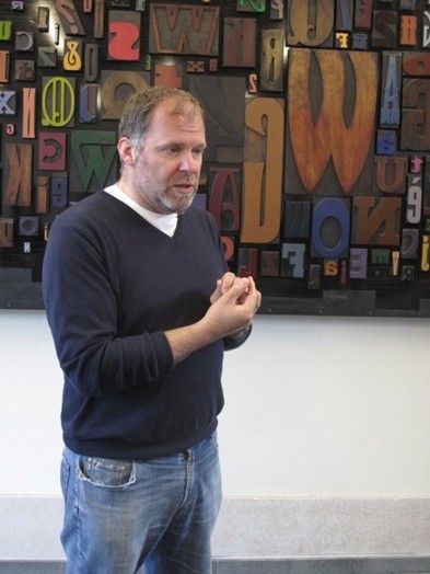 A photo of a man talking and behind him a board full of different color fonts carved in wood.