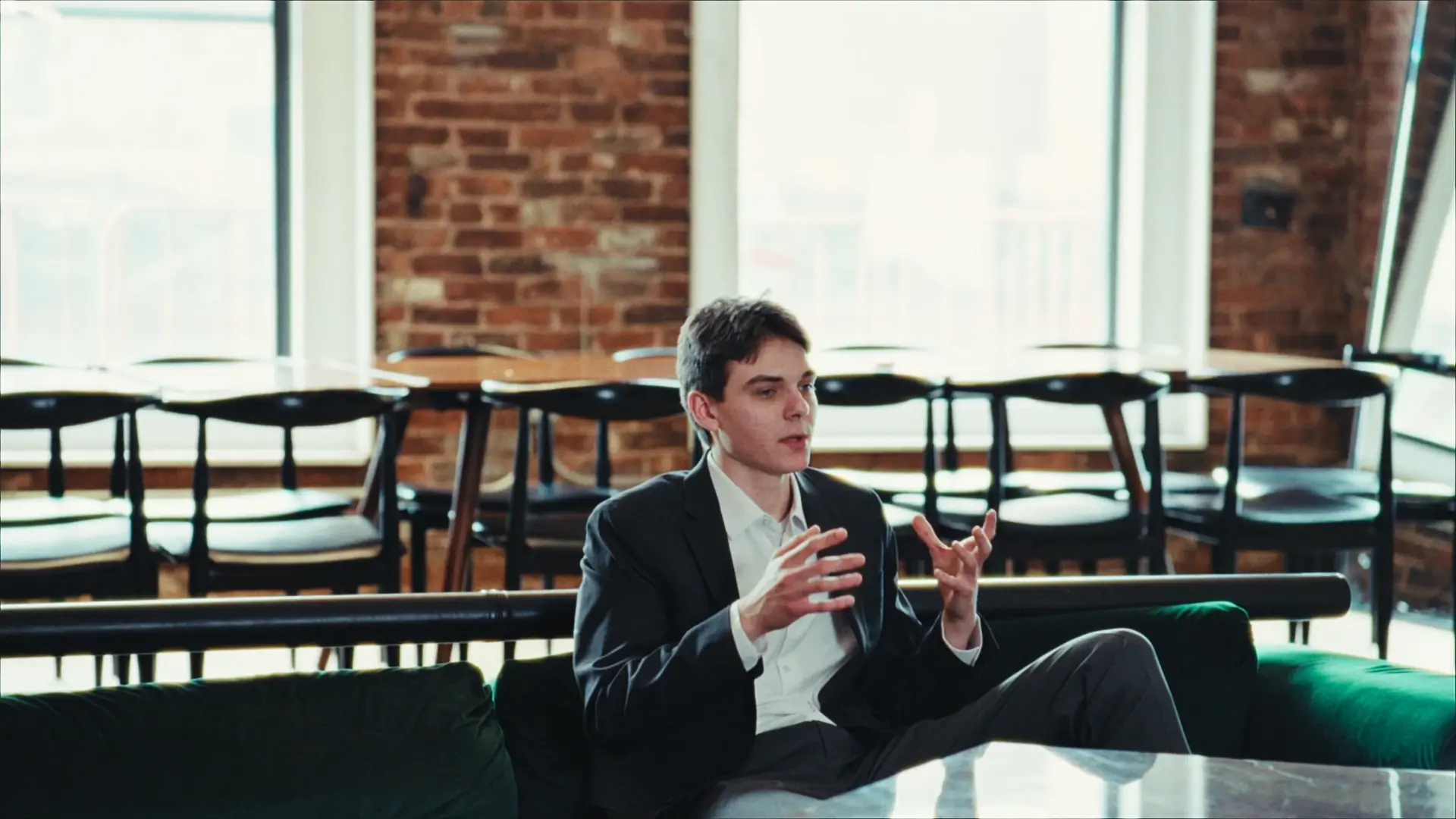 A man in a suit speaking at a table in a loft with a brick wall