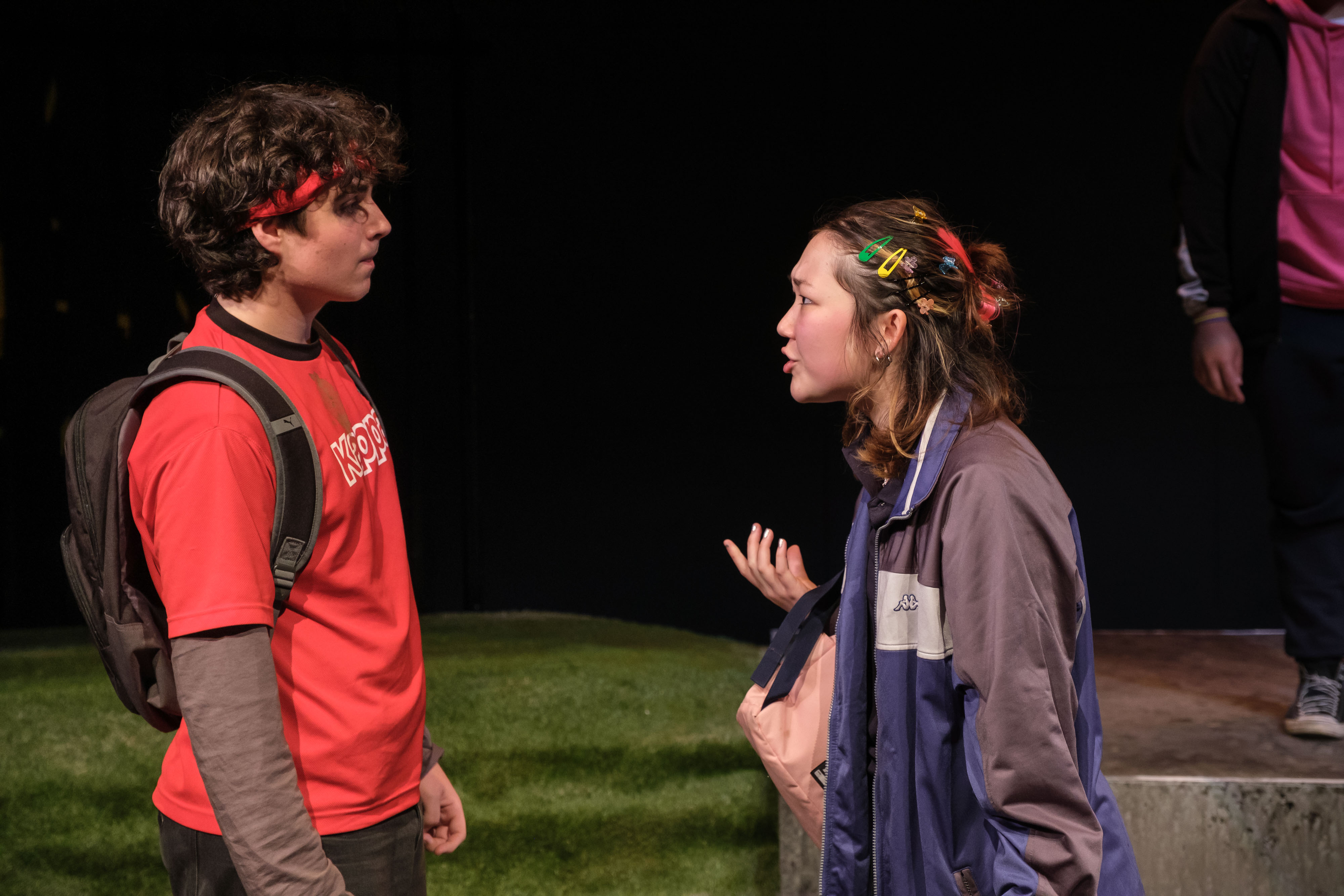 Two teenage performers stand face-to-face in a tense conversation. One wears a red T-shirt and backpack, while the other, wearing a school jacket covered in colourful hair clips, gestures expressively as if confronting a classmate.