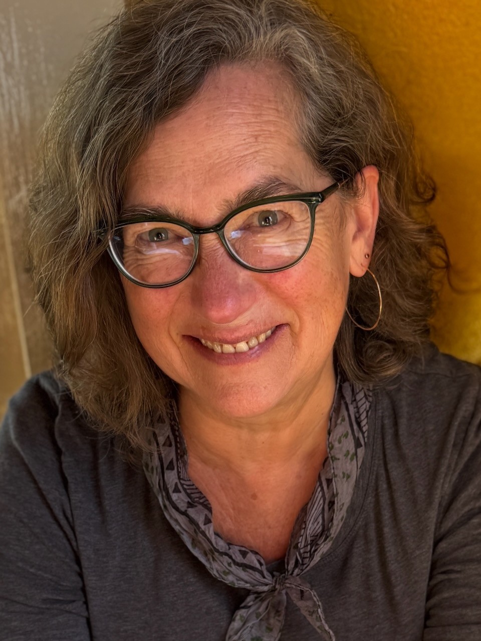 Cate, smiling at the camera, wearing glasses, earrings, and a scarf, in a sunny room