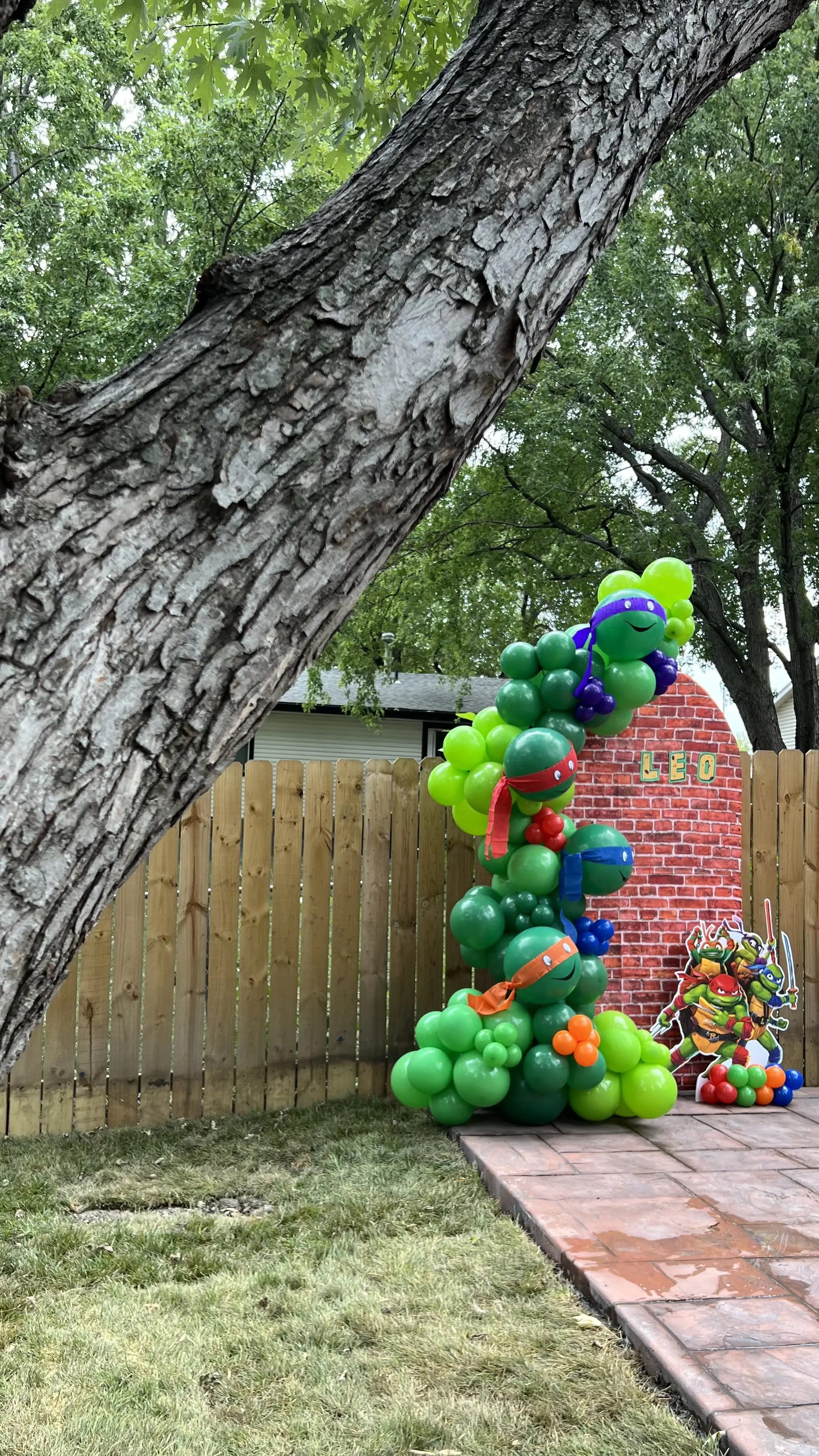 Ninja Turtles-themed balloon garland with green and blue balloons arranged in an organic arch against a brick wall backdrop. Perfect for kids who love superhero adventures. Susa’s Sweets & Balloons.