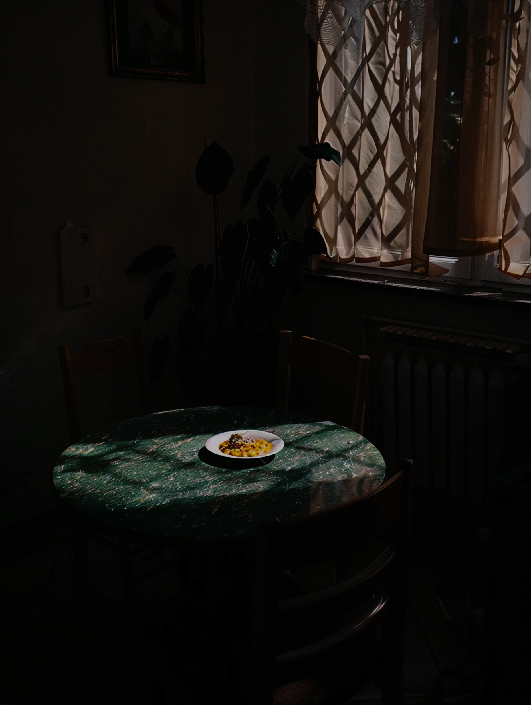 Dimly lit room with sunlight filtering through curtains, casting shadows on a green marble table with a single plate of pasta in the center.
