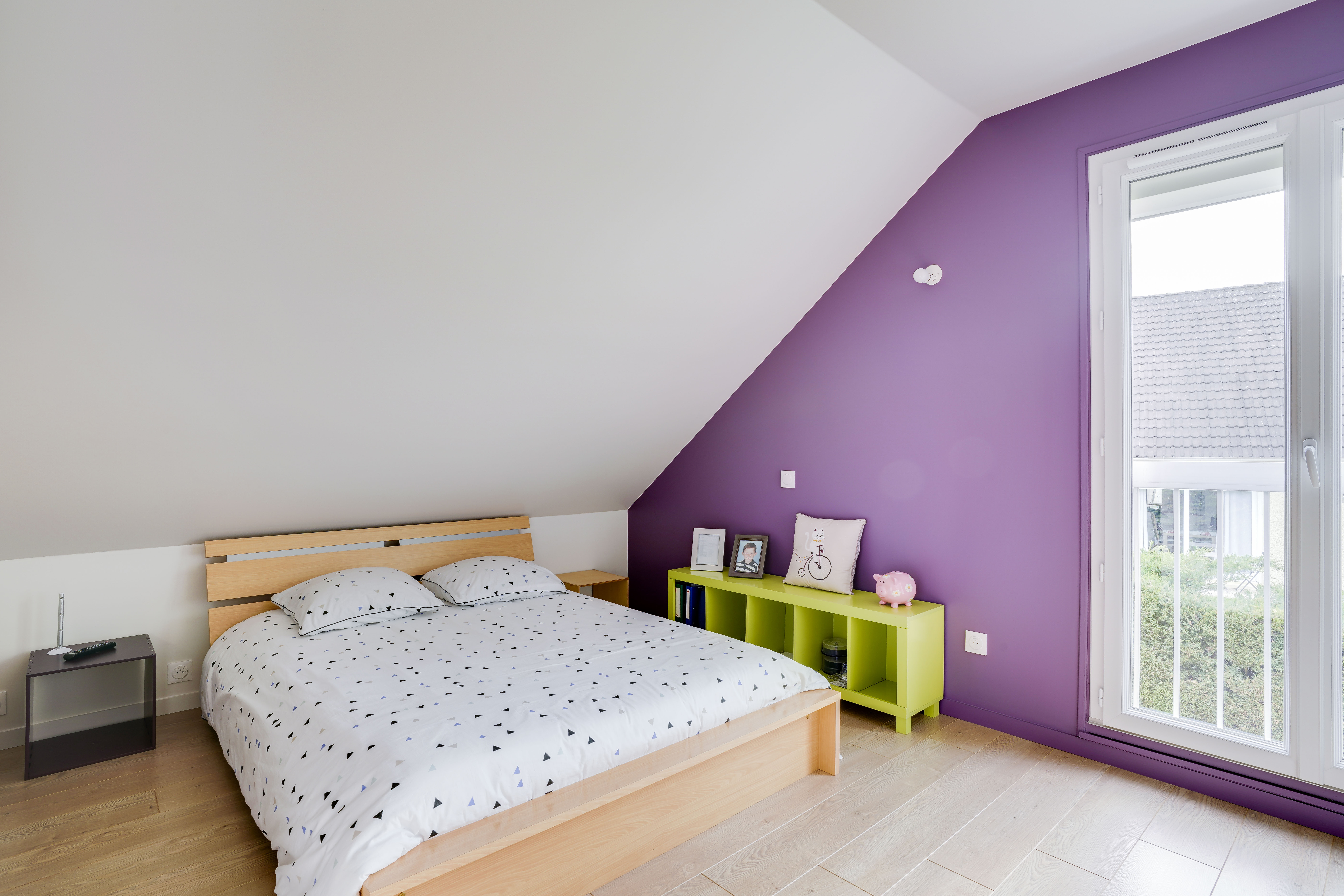 Rénovation chambre mansardée 77 avec plafond incliné blanc, lit bois, étagère verte et mur accent violet, transformation 3 pièces.