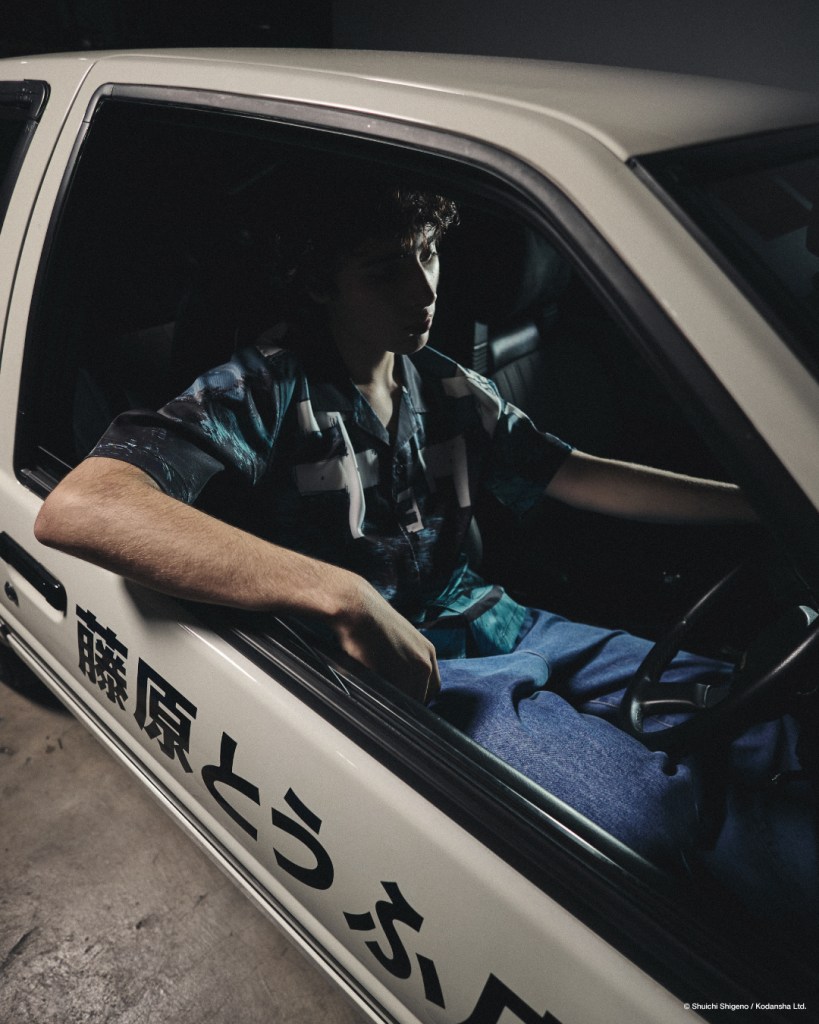 A young man sitting inside a white car, wearing a patterned shirt, with a contemplative expression. The car features Japanese text on the side, reflecting a street racing culture.