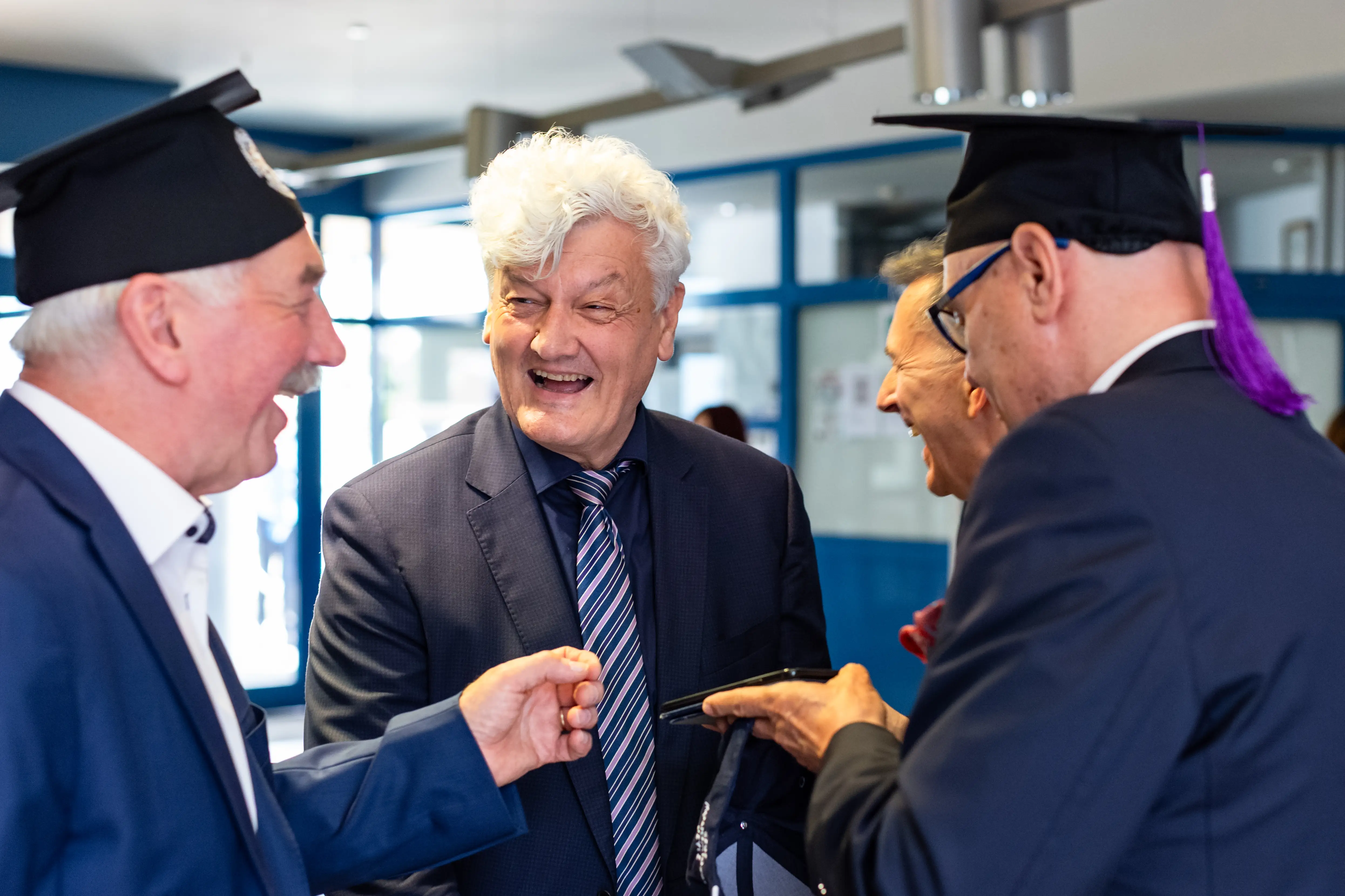 Czterej mężczyźni rozmawiają w spokojnej atmosferze z uśmiechami, każdy z nich w garniturach i charakterystycznych czapkach, typowych dla uroczystości akademickich, na tle jasnej przestrzeni wnętrza.