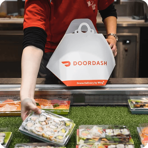 A person in a red uniform places a packaged sushi tray on a counter beside a branded DoorDash delivery container, highlighting food delivery services.